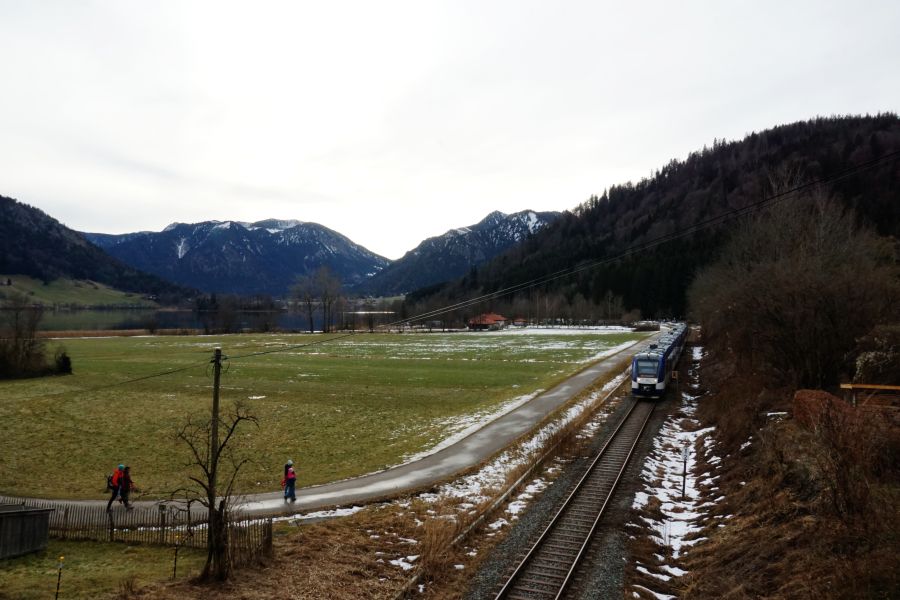 Eine Landschaft in Bayern, mit grünen Wiesen, schneebedeckten Bergen im Hintergrund und einem Zug, der entlang der Gleise fährt. Zwei Wanderer gehen einen Weg entlang.