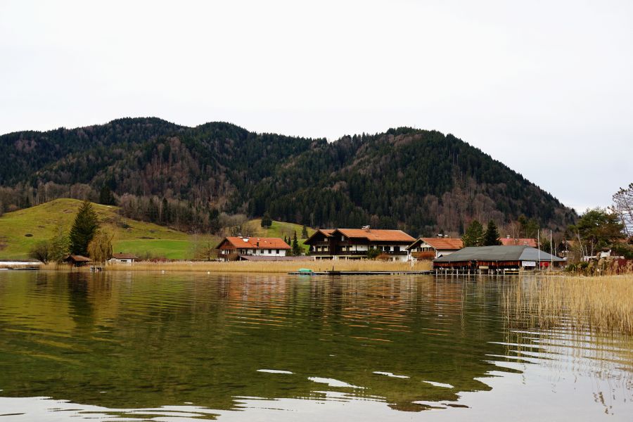 An der Uferpromenade des Schliersees zeigt sich eine malerische Aussicht auf die sanften Hügel und Häuser im Hintergrund, umgeben von bewaldeten Bergen, an einem bewölkten Tag.