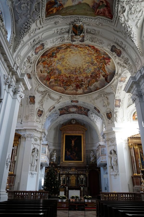 Innenansicht einer Kirche mit einem kunstvoll bemalten Deckenfresko und einer majestätischen Altaranlage, die mit reich verzierten Säulen und Statuen ergänzt wird. Im Vordergrund stehen Holzbänke.