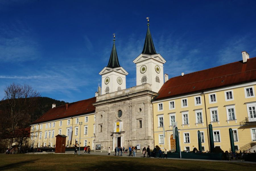 Die Vorderansicht eines historischen Gebäudes mit zwei spitzdachigen Türmen und einer zentralen Eingangstür. Menschen stehen vor dem Gebäude, das von einem blauen Himmel und Bäumen umgeben ist.