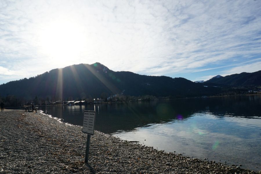 Sonnenstrahlen spiegeln sich im Wasser des Schliersees, im Hintergrund erheben sich die Berge unter einem klaren Himmel.