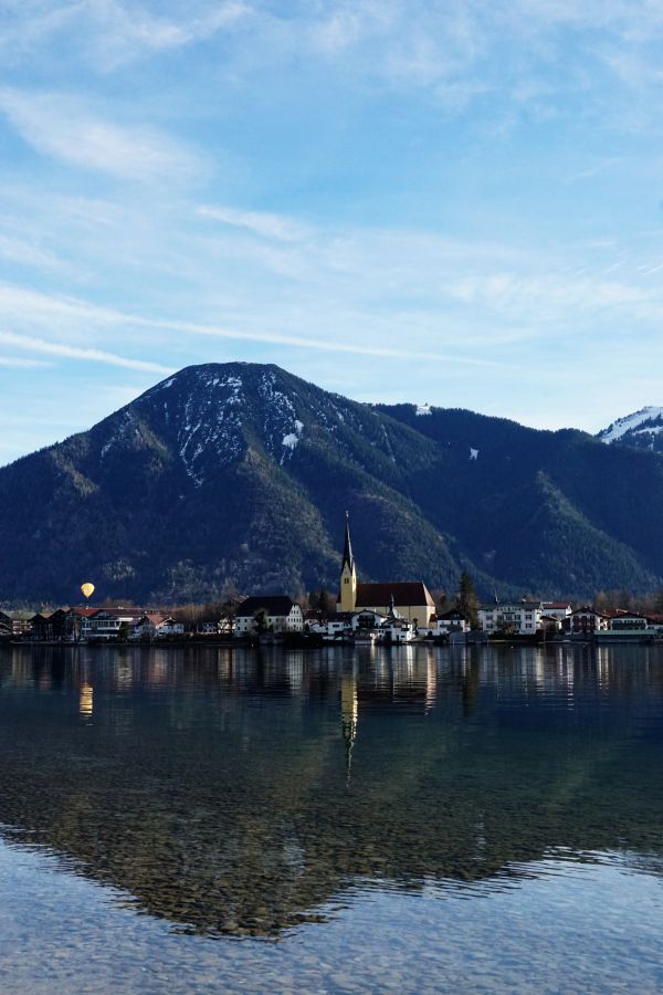 Blick auf den Schliersee mit reflektierenden Bergen und einem kleinen Ort mit Kirche im Vordergrund