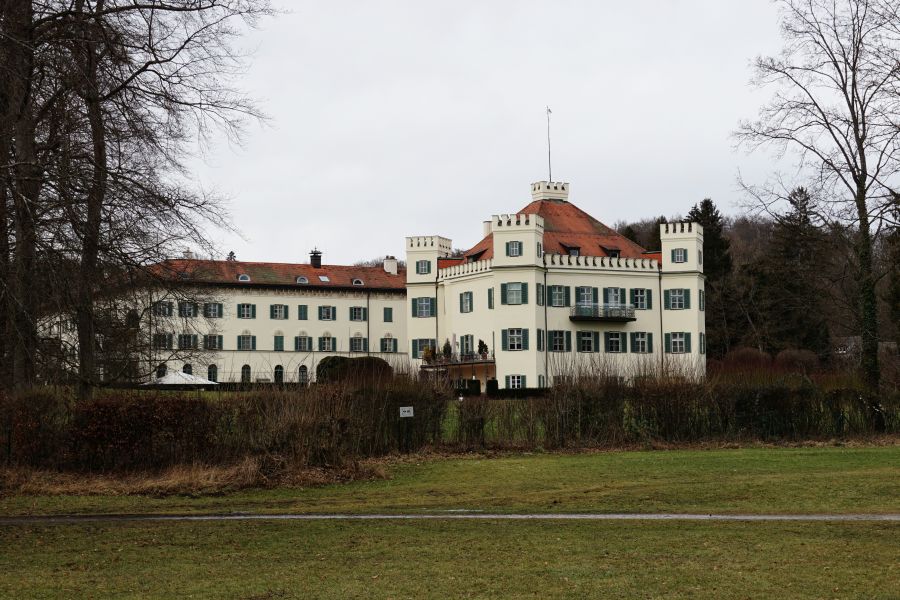 Ein historisches Gebäude mit einem roten Dach und weißen Wänden, umgeben von Bäumen und Rasen. Der Himmel ist grau und es ist ein kühler Tag.