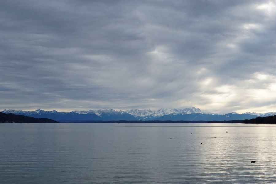 Der Starnberger See mit schneebedeckten Bergen im Hintergrund und einem bewölkten Himmel.