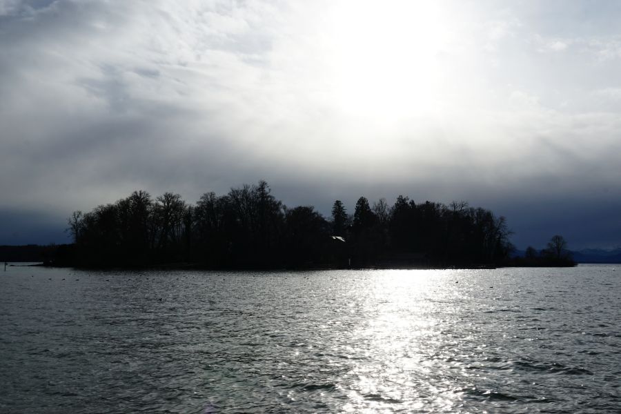Eine ruhige Seelandschaft mit Bäumen am Ufer, während die Sonne hinter dunklen Wolken aufgeht und das Wasser in sanften Wellen schimmert.