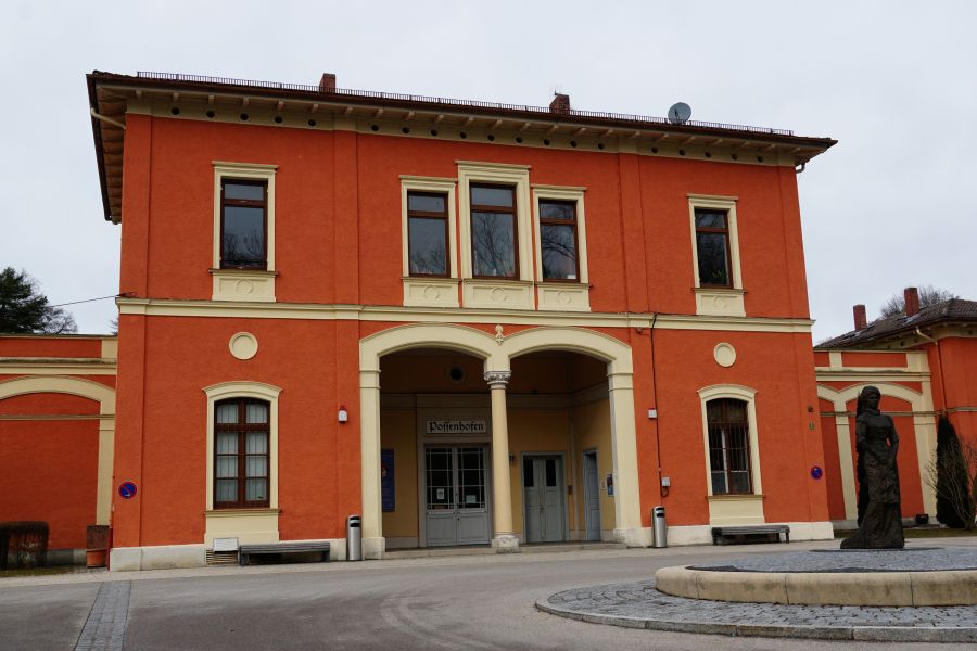 Das orangefarbene Gebäude des Bahnhofs in Possenhofen mit Landungsplattform und einer Statue im Vordergrund.