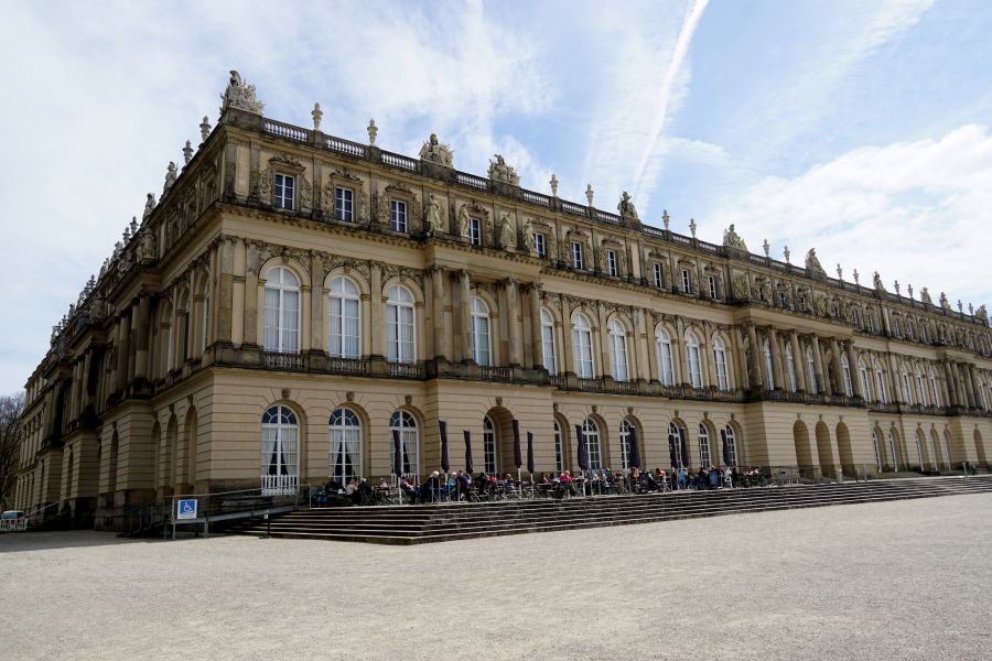 Ansicht des Neuen Schlosses Herrenchiemsee mit mehreren Fenstern und Statuen auf dem Dach, umgeben von Besuchern und einem hellen Himmel.