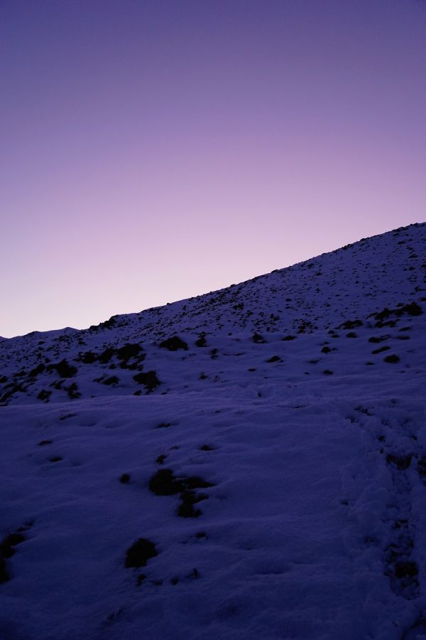 Ein schneebedeckter Hang unter einem violetten Abendhimmel, mit sanften Wellen und Schatten, die die Landschaft formen.