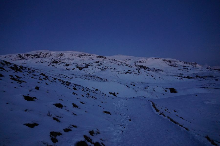 Schneebedeckte Landschaft bei Dämmerung mit sanften Hügeln und Bergen im Hintergrund.