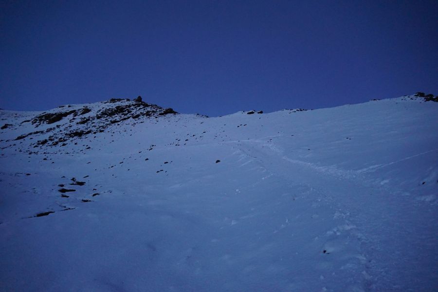 Ein schneebedeckter Berg mit grauen Steinen im Vordergrund unter einem dunklen Himmel.