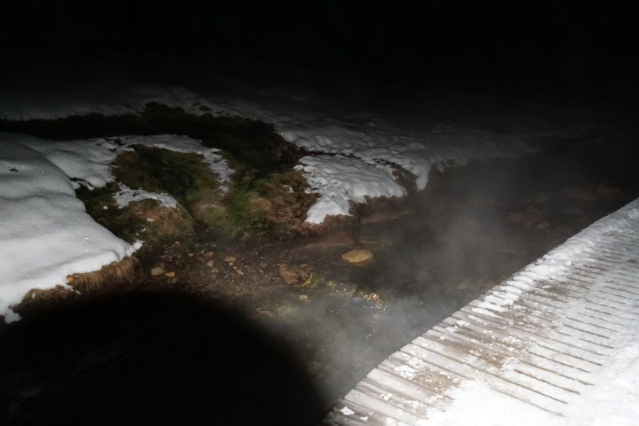 Ein dampfender Fluss fließt durch eine verschneite Landschaft in der Dunkelheit, mit Schnee und nassen Stellen an den Ufern.