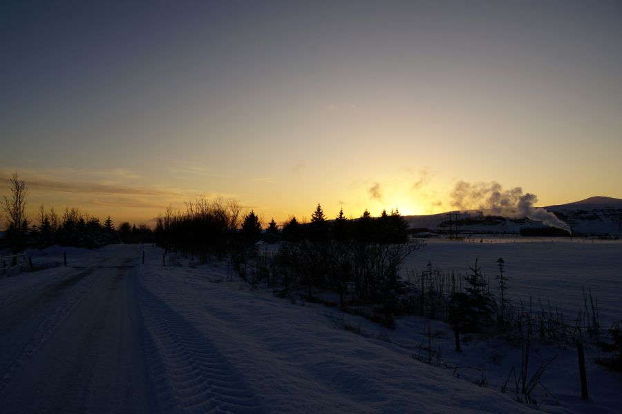 Winterliche Landschaft mit einem schneebedeckten Weg, Bäumen und einem Sonnenuntergang im Hintergrund. Dampf steigt aus einer Quelle auf und der Himmel leuchtet in warmen Farben.