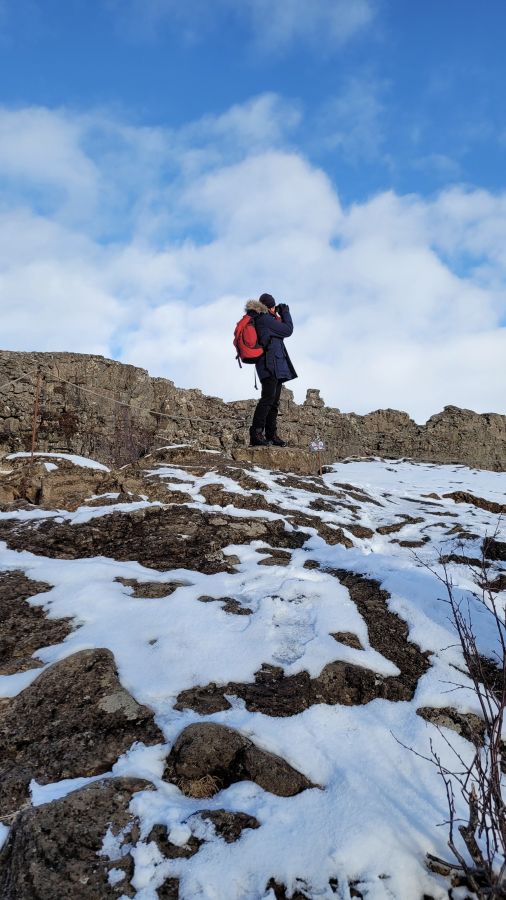 Ein Reisender steht auf einem schneebedeckten Felsen und fotografiert die Landschaft unter einem klaren blauen Himmel.