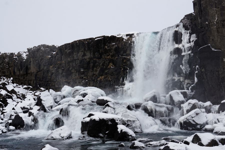 Ein gefrorener Wasserfall in einer isländischen Landschaft, umgeben von schneebedeckten Felsen und eisigen Strukturen, unter einem grauen Himmel.