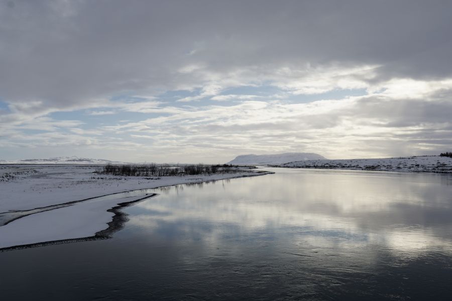 Eine ruhige Winterlandschaft in Island mit einem schneebedeckten Ufer und einem stillen, reflektierenden Fluss unter einem bewölkten Himmel.