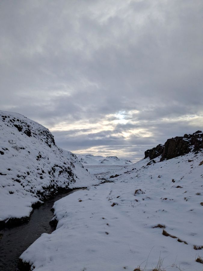 Eine verschneite Landschaft mit einem schmalen Fluss, der zwischen den schneebedeckten Hügeln fließt. Der Himmel ist bewölkt und zeigt einen schwachen Lichtschein.