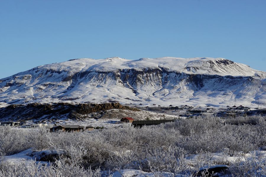 Panorama eines verschneiten Berges in Island mit klar blauem Himmel, im Vordergrund sind schneebedeckte Wiesen und einige kleine Gebäude zu sehen.