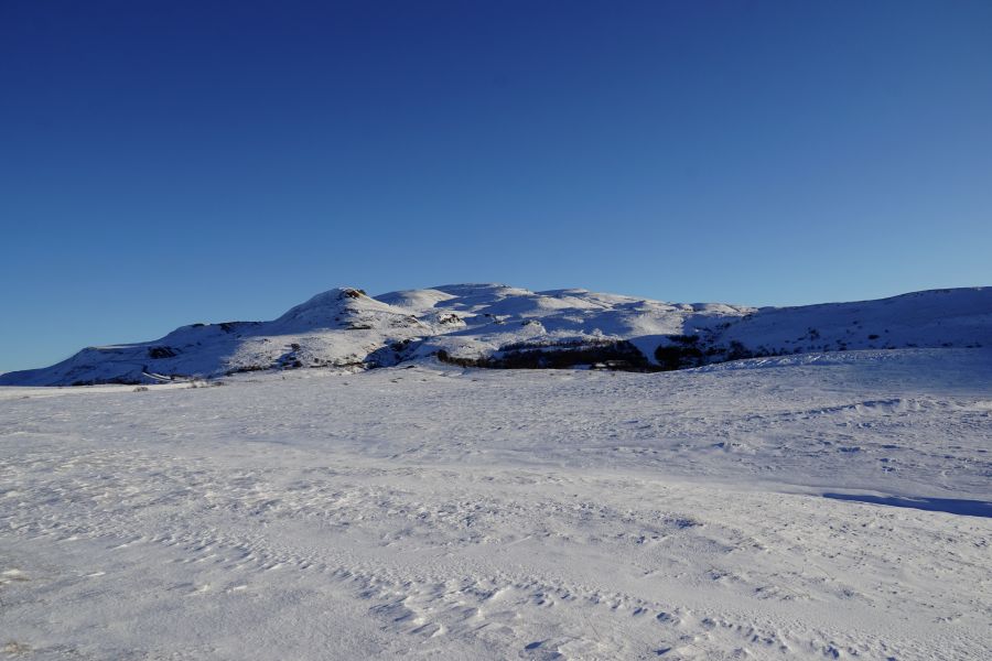 Eine weite, schneebedeckte Landschaft mit einem Hügel im Hintergrund unter einem klaren blauen Himmel.
