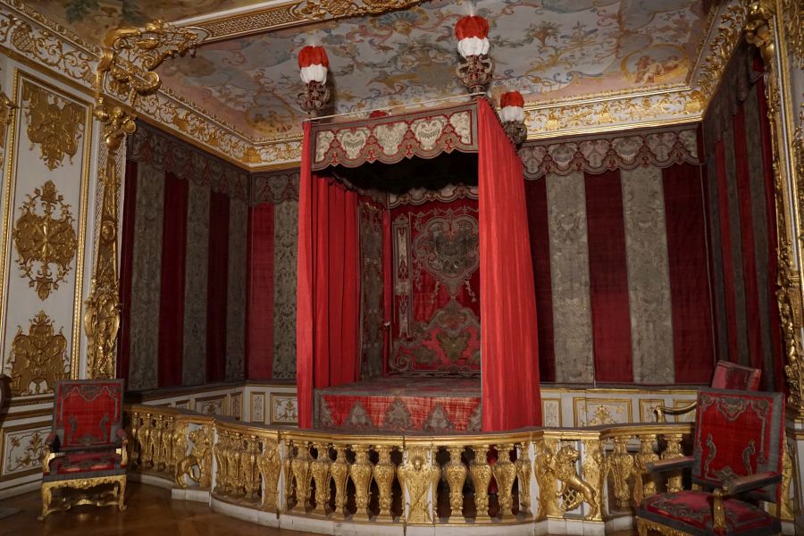 Detailansicht eines barocken Schlafzimmers im Schloss Schleißheim, mit rotem Vorhang, goldenen Verzierungen und prächtigem deckenfresko.