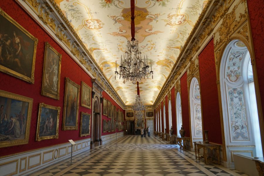 Ein prunkvoller Saal in der barocken Schlossanlage Schleißheim, mit opulenter Deckenmalerei, roten Wänden und goldverzierten Rahmen um historische Gemälde.