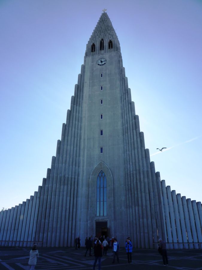 Die Hallgrímskirkja in Reykjavík, eine markante Betonarchitektur mit einem hohen Turm und einem klaren blauen Himmel im Hintergrund, umgeben von Menschen.