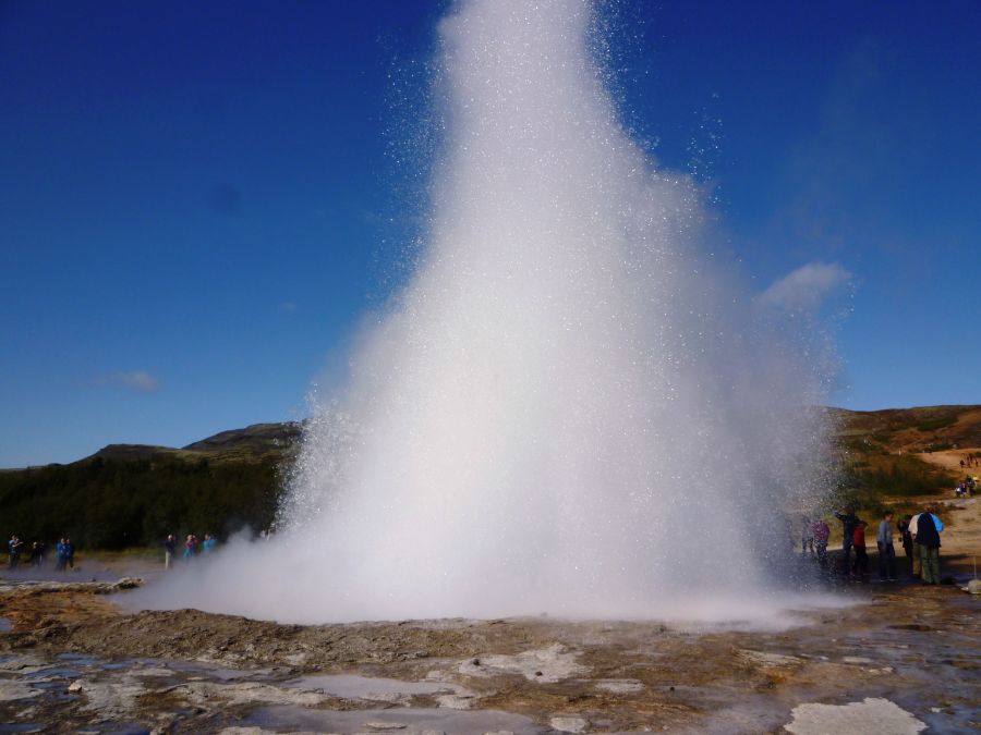 Ein Geysir, der Wasser in die Luft schleudert, umgeben von einer klaren blauen sky. Im Hintergrund sind Menschen zu sehen, die das Naturschauspiel beobachten.