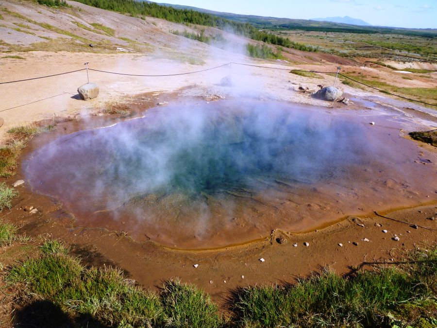 Ein dampfendes heißes Quellbecken in einer grauen Steinlandschaft, umgeben von grünem Gras und sanften Hügeln. Der Dampf steigt ruhig aus dem Wasser auf und vermittelt eine mystische Atmosphäre.