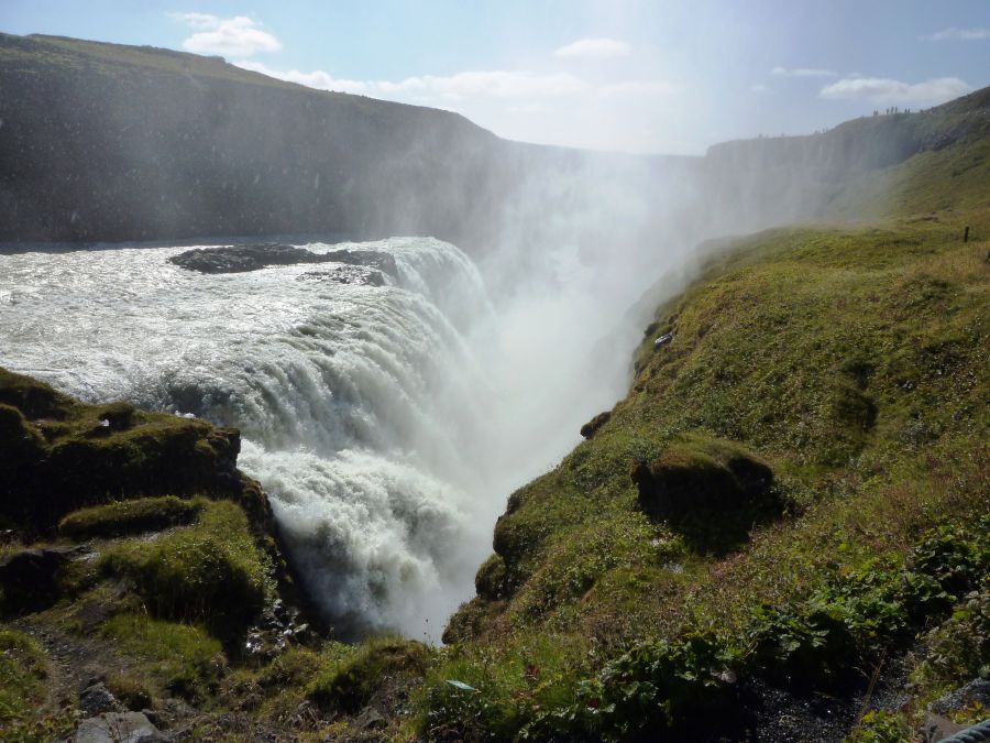 Ein beeindruckender Wasserfall stürzt mit kraftvollem Wasser über eine Klippe, umgeben von grünen Wiesen und steilen Felsen unter einem klaren Himmel.
