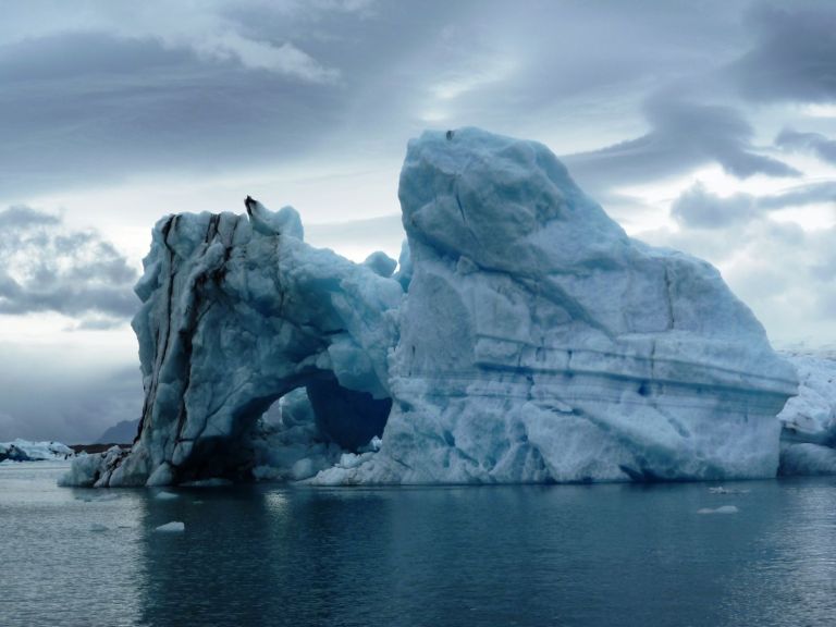 Eine beeindruckende Eisscholle mit einer durchbrochenen Struktur, die im ruhigen Wasser einer Gletscherlagune schwimmt, unter einem bewölkten Himmel.