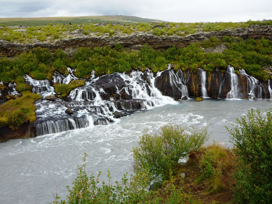 Ein Wasserfall fließt über schwarze Felsen in einen grauen Fluss, umgeben von grüner Vegetation und hügeliger Landschaft.