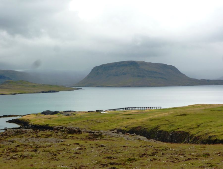 Eine ruhige Landschaft mit einem See im Vordergrund, umgeben von sanften Hügeln und bewölktem Himmel.