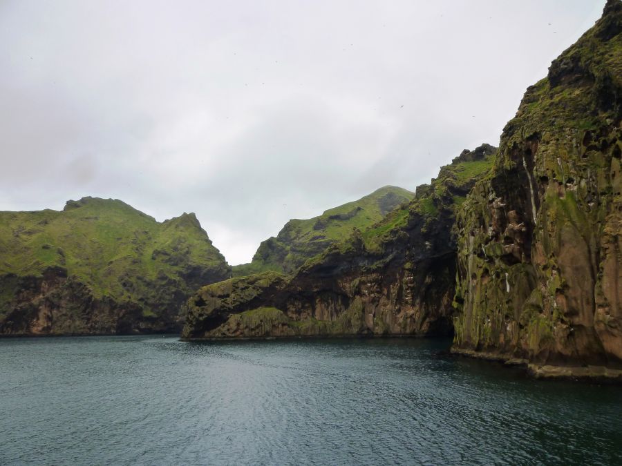 Grüne Klippen und Berge umgeben eine ruhige, klare Wasserfläche, die im Vordergrund zu sehen ist. Der Himmel ist bewölkt, was der Szenerie eine mystische Atmosphäre verleiht.