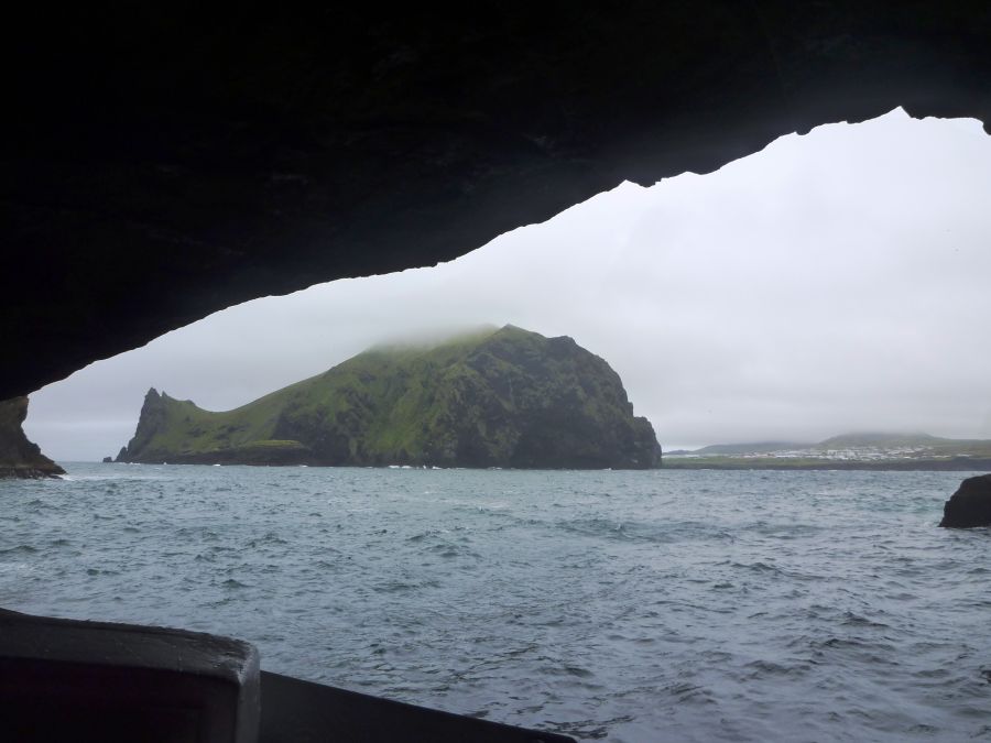 Blick aus einer Höhle auf eine grüne Insel und das ruhige Meer bei bewölktem Himmel.