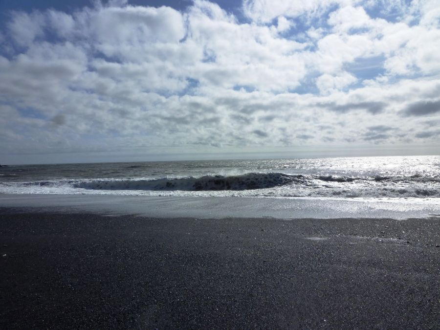 Schwarzer Sandstrand mit Wellengang und bewölktem Himmel, reflektiert von der Wasseroberfläche.