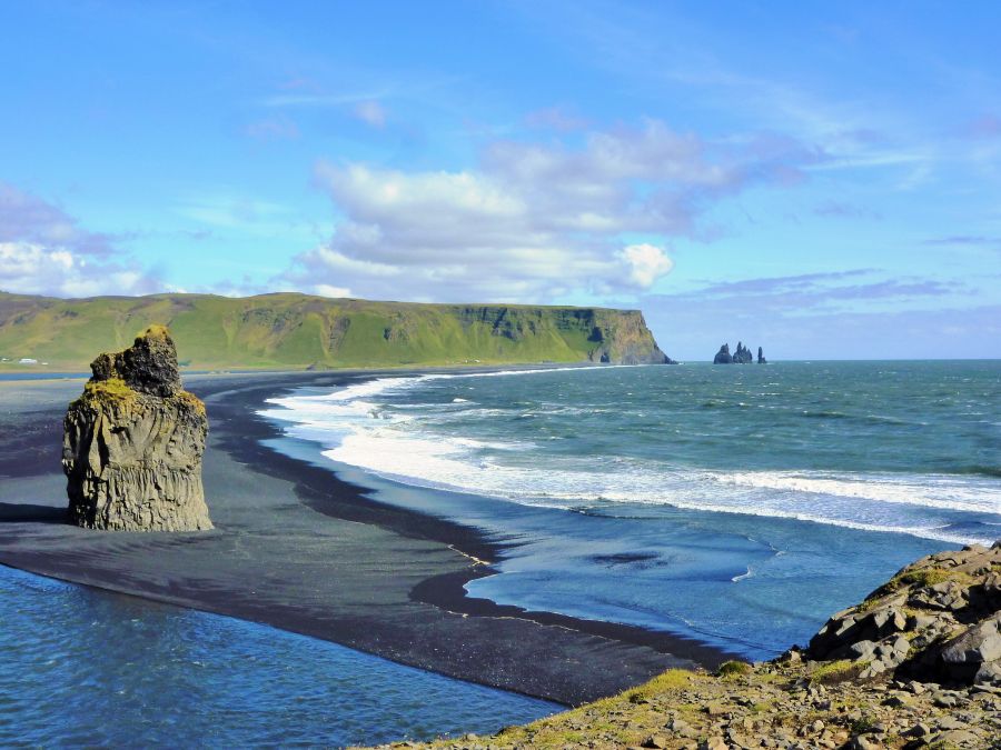 Ein Blick auf die Küste von Island, mit schwarzem Sandstrand, klarem Wasser und dramatischen Felsformationen im Hintergrund unter einem blauen Himmel.