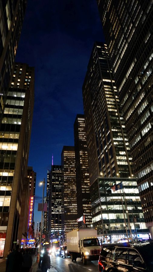 Blick auf eine belebte Straße in New York City bei Nacht, umgeben von hohen Bürogebäuden, mit bunten Lichtern und Verkehr.