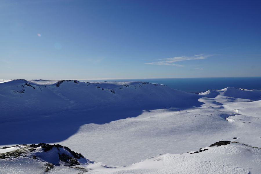 Eine weite, verschneite Landschaft mit sanften Hügeln und einem klaren blauen Himmel über dem Meer.