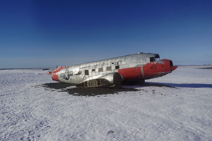 Ein beschädigtes US-amerikanisches Flugzeugwrack einer DC-3 liegt auf einem schneebedeckten Feld in Island unter klarem Himmel.