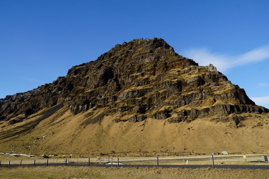 Ein steiler, rauer Hügel mit schroffem Fels und grün-brauner Vegetation unter einem wolkenlosen blauen Himmel.