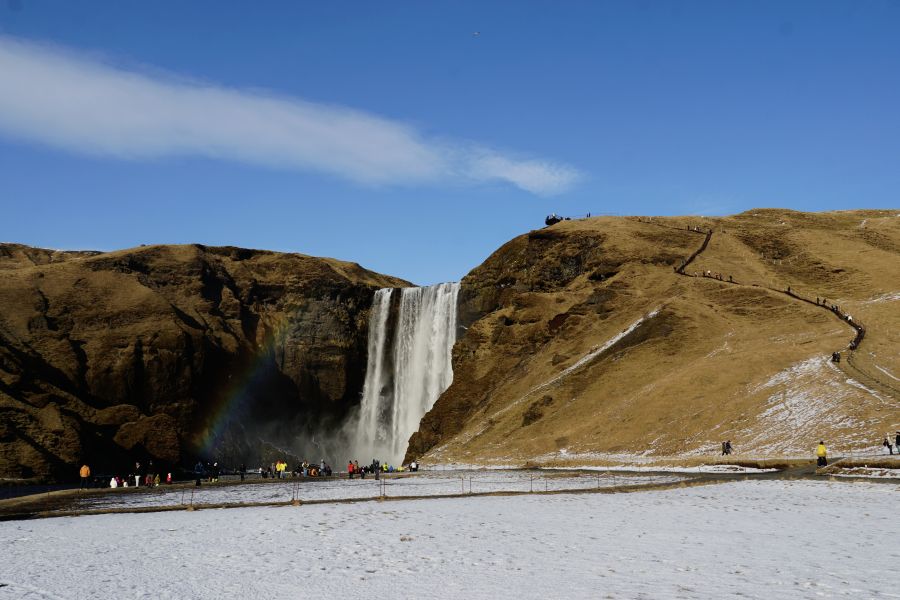 Der Skógafoss Wasserfall in Island, umgeben von schneebedeckten Wiesen und hügeliger Landschaft, mit einem Regenbogen, der sich im Wasser bildet. Touristen stehen am Fuß des Wasserfalls und genießen die Aussicht unter einem klaren, blauen Himmel.