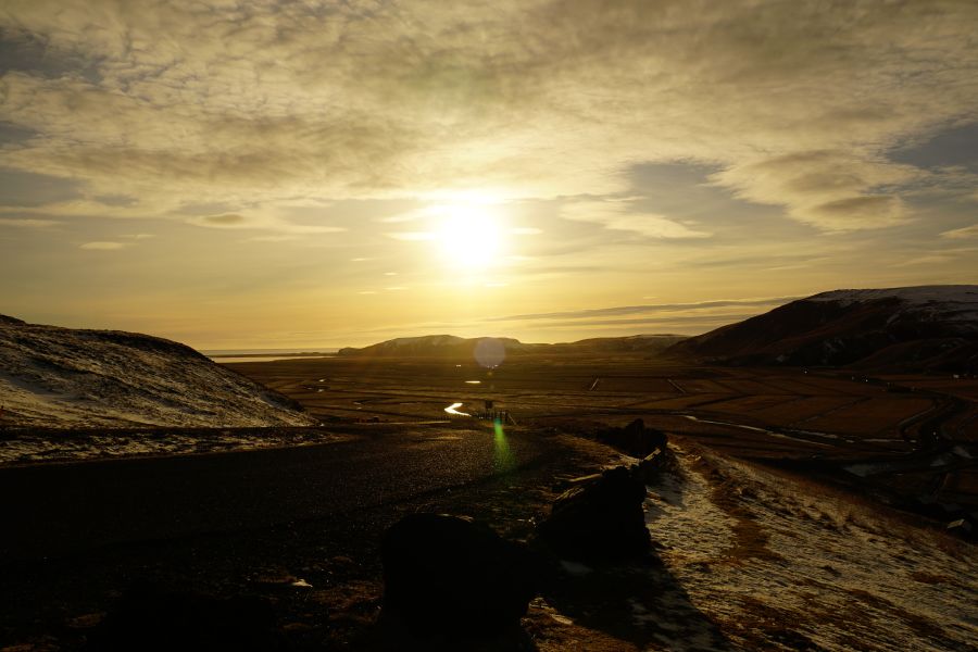 Sonnenuntergang über einer weiten, verschneiten Landschaft in Island, mit sanften Hügeln und einer ruhigen Straße im Vordergrund.