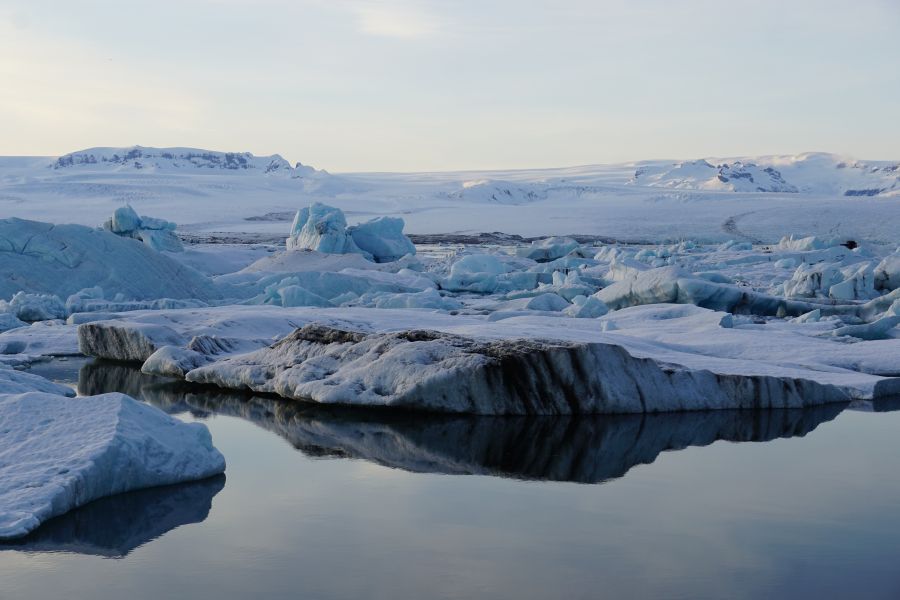 Eisschollen schwimmen in einem Gletschersee, umgeben von schneebedeckten Bergen und der ruhigen Reflektion des Himmels auf dem Wasser.
