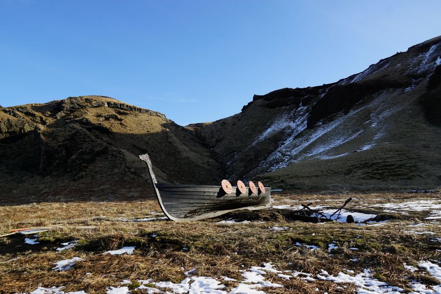 Eine alte Wikingerschiffsnachbildung liegt auf einer Wiese, umgeben von sanften Hügeln und Schnee.