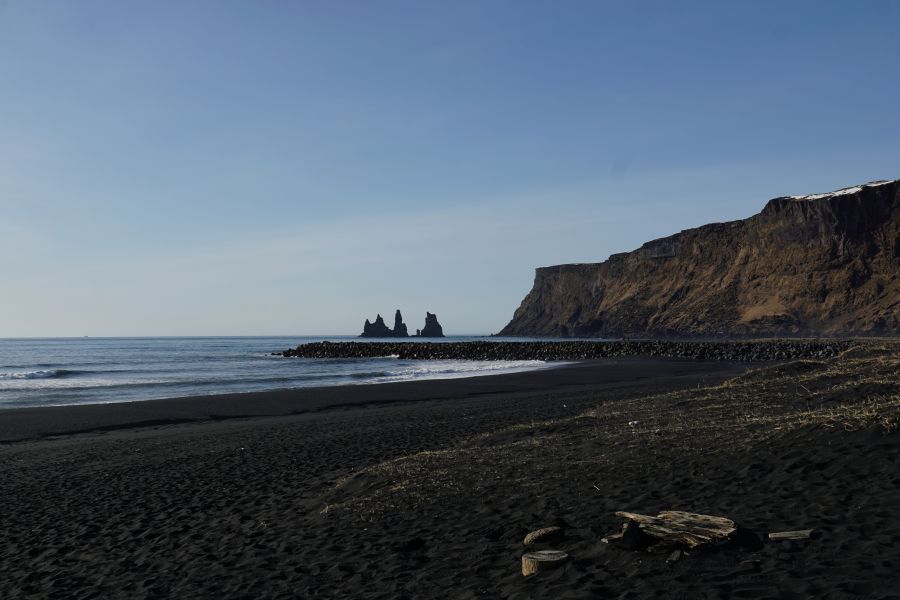 Blick auf den schwarzen Sandstrand in Vík í Mýrdal mit den charakteristischen Felsformationen Reynisdrangar im Hintergrund und einer steilen Küstenlinie.