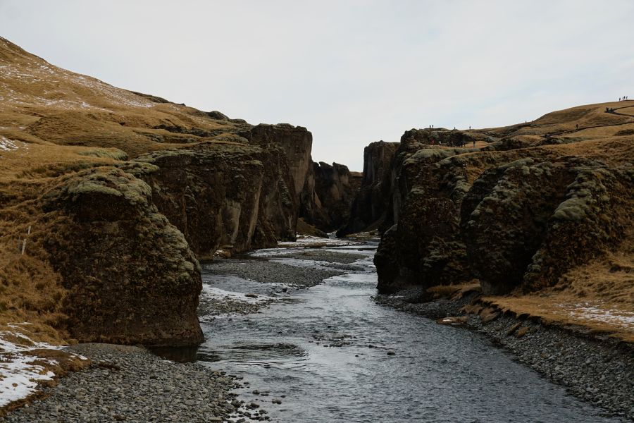 Eine beeindruckende Flussschlucht mit steilen Felswänden, über die Wasser fließt, umgeben von sanften Hügeln mit teilweise schneebedecktem Gras.