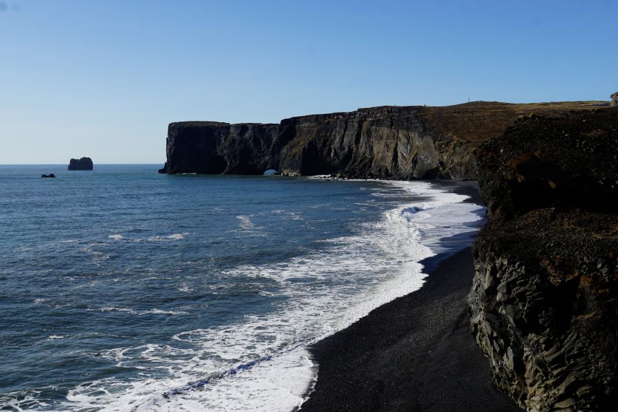 Blick auf die Küste mit dramatischen Klippen und dem Ozean, sanfte Wellen brechen am schwarzen Sandstrand.