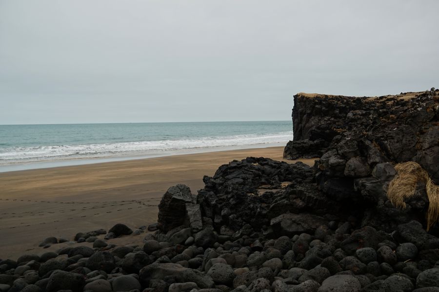 Ein ruhiger Strand mit schwarzem Vulkangestein, grauem Himmel und sanften Wellen des Ozeans.