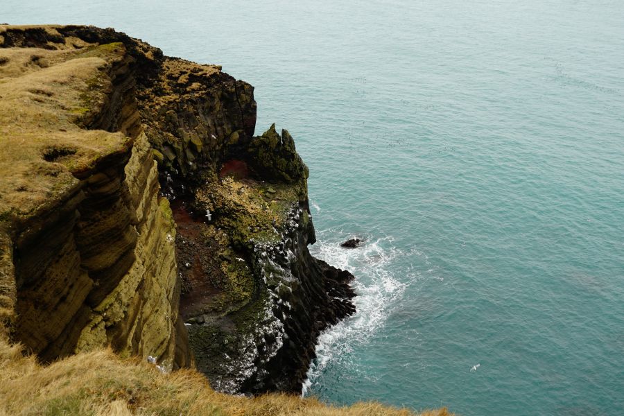 Eine steile Klippe, die ins ruhige türkisfarbene Wasser des Ozeans abfällt. Der Rand der Klippe ist mit trockenem Gras bewachsen, während das Wasser sanft gegen die Felsen plätschert.