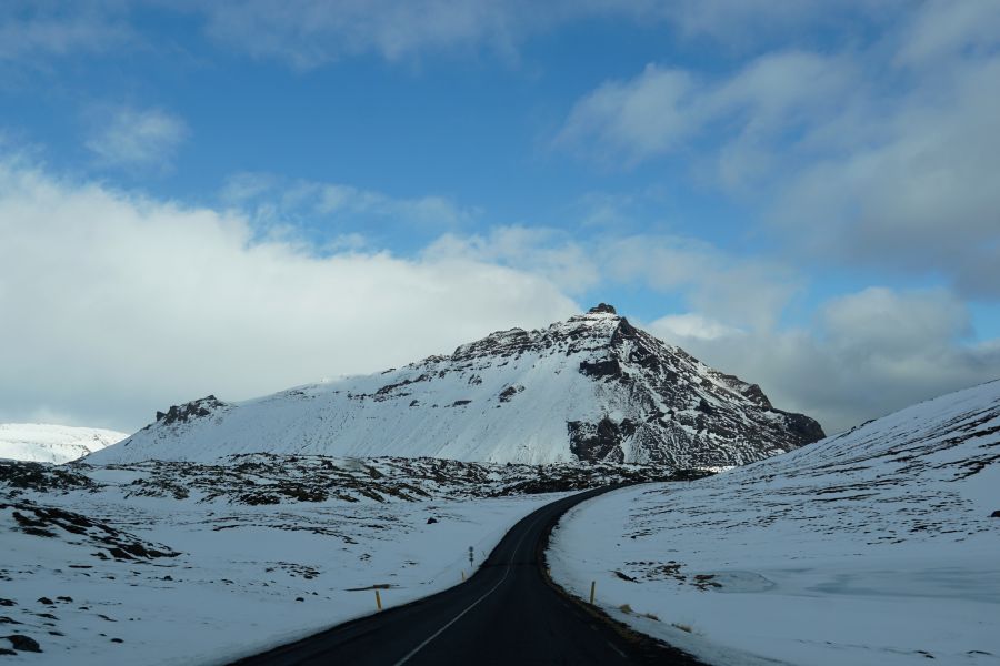 Ein schneebedeckter Berg mit einer kurvigen Straße im Vordergrund, unter blauem Himmel mit einigen Wolken.