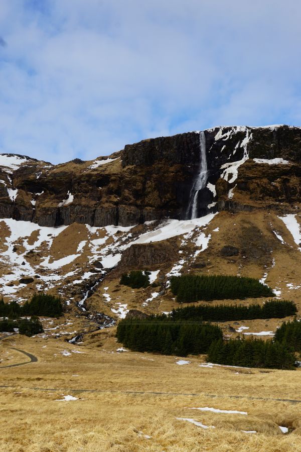 Ein Wasserfall fließt über eine hohe Felswand, umgeben von schneebedeckten Bergen und trockener, brauner Landschaft mit einigen grünen Baumgruppen im Vordergrund.